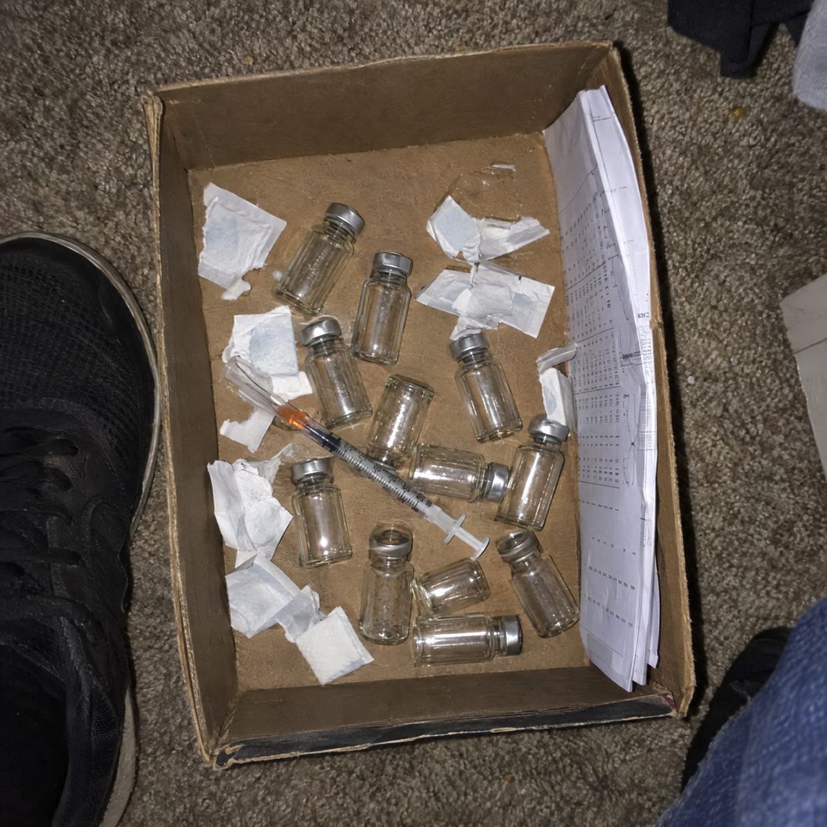 Shoebox on a closet floor full of small vials and a syringe