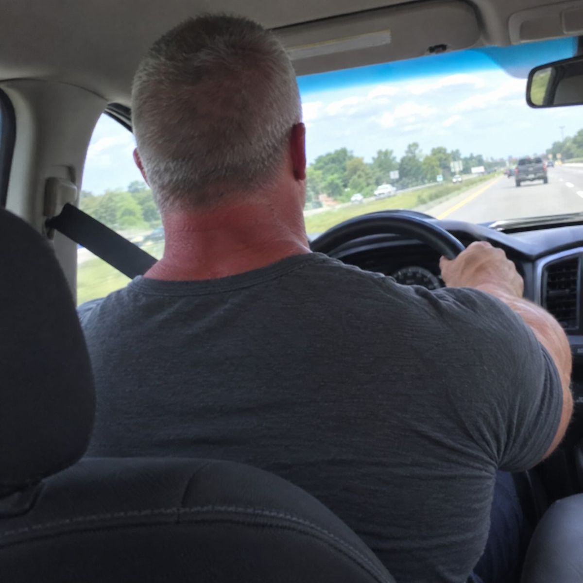 Man from behind in the driver's seat, traps and shoulders stretching a grey t-shirt